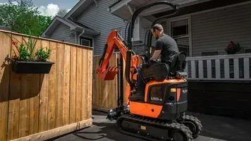 Man operating an orange Kubota mini-excavator near a wooden fence and house.