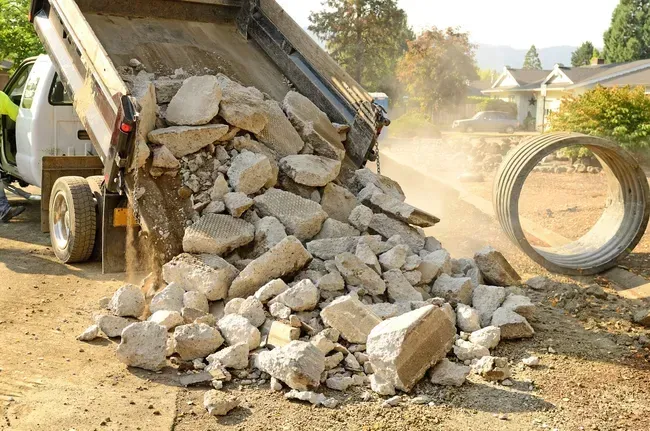 Dump truck unloading broken concrete on a dirt road next to a metal drainage pipe.