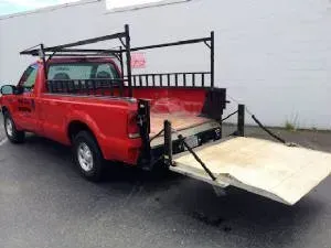 Red pickup truck with a wooden platform ramp lowered. It has a ladder rack.