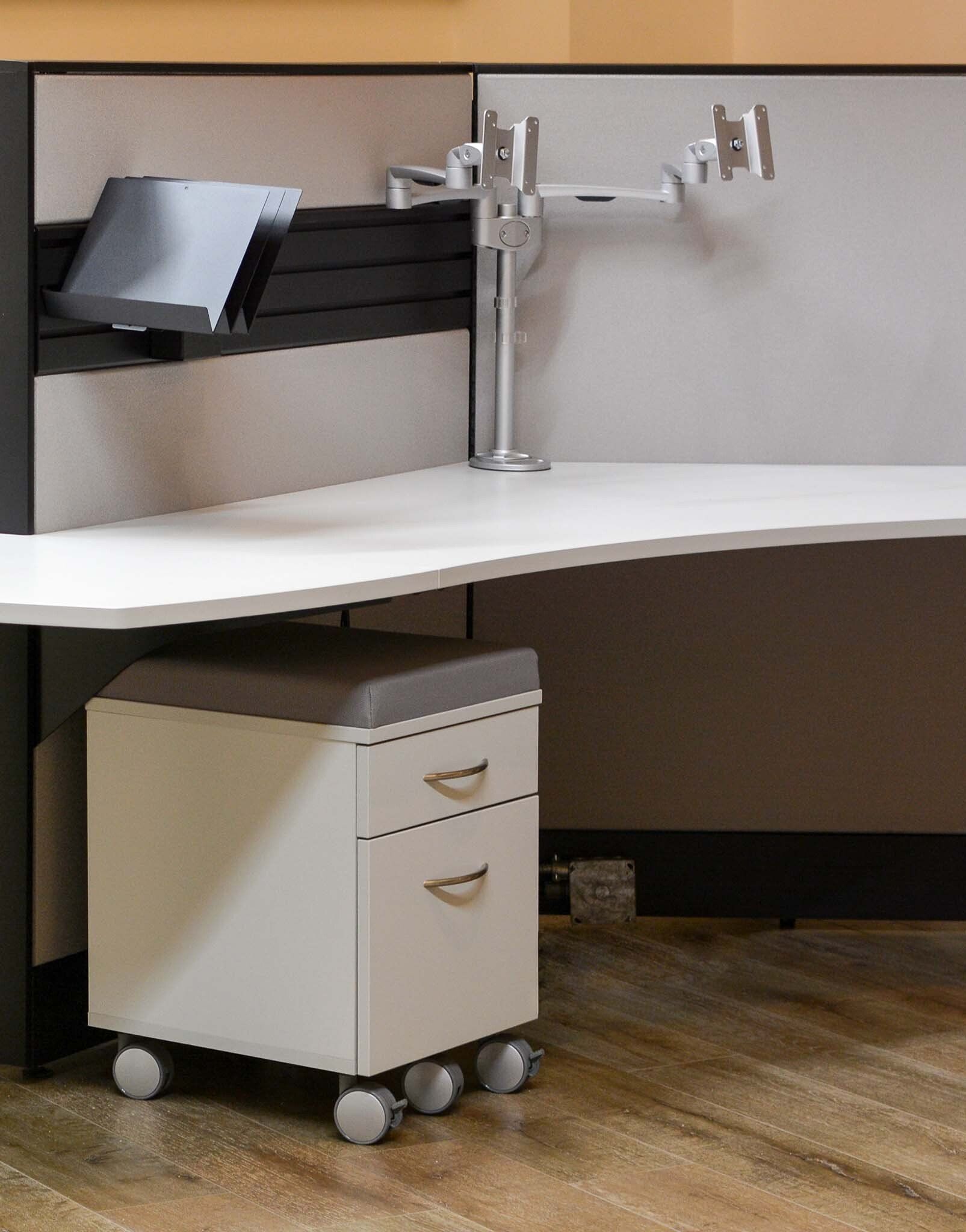 a cubicle with two monitor arms and a filing cabinet