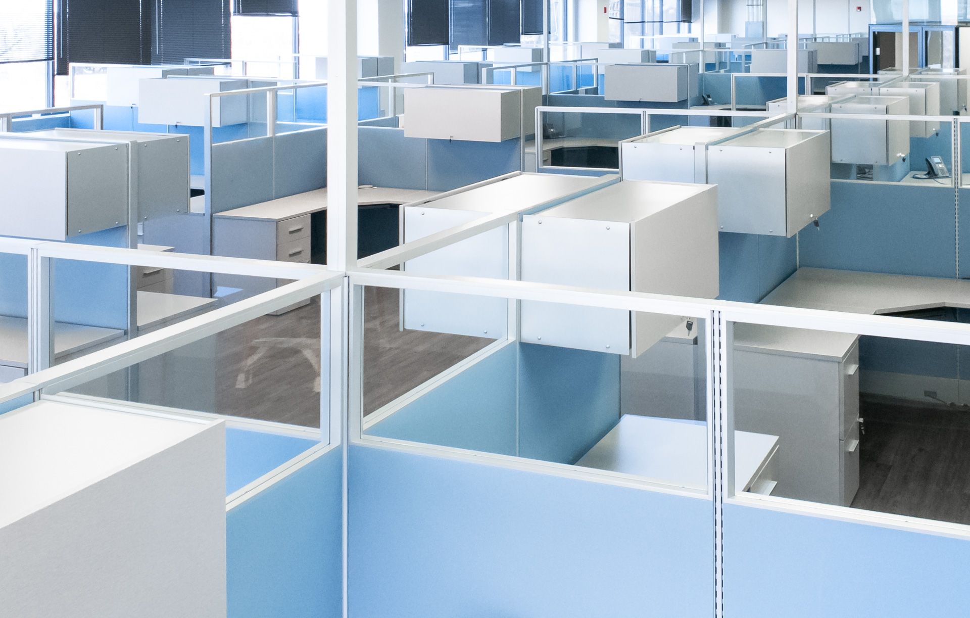 Large room with rows of workstations with clear windows, white storage cabinets and light blue fabric panels
