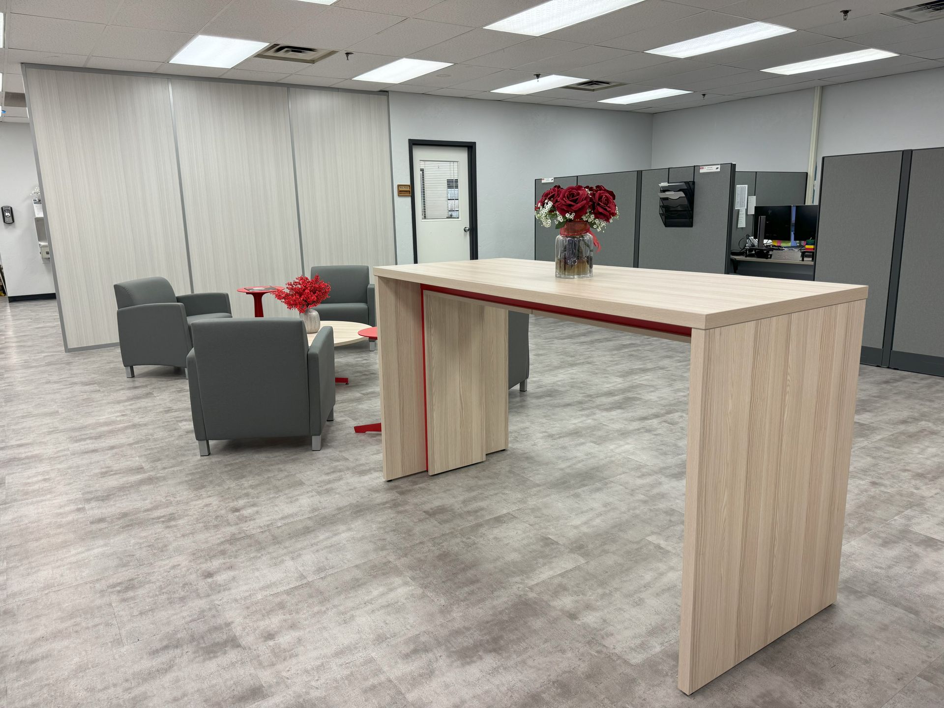 An office waiting area with a light wood table, gray chairs, red accents, and a vase of flowers.