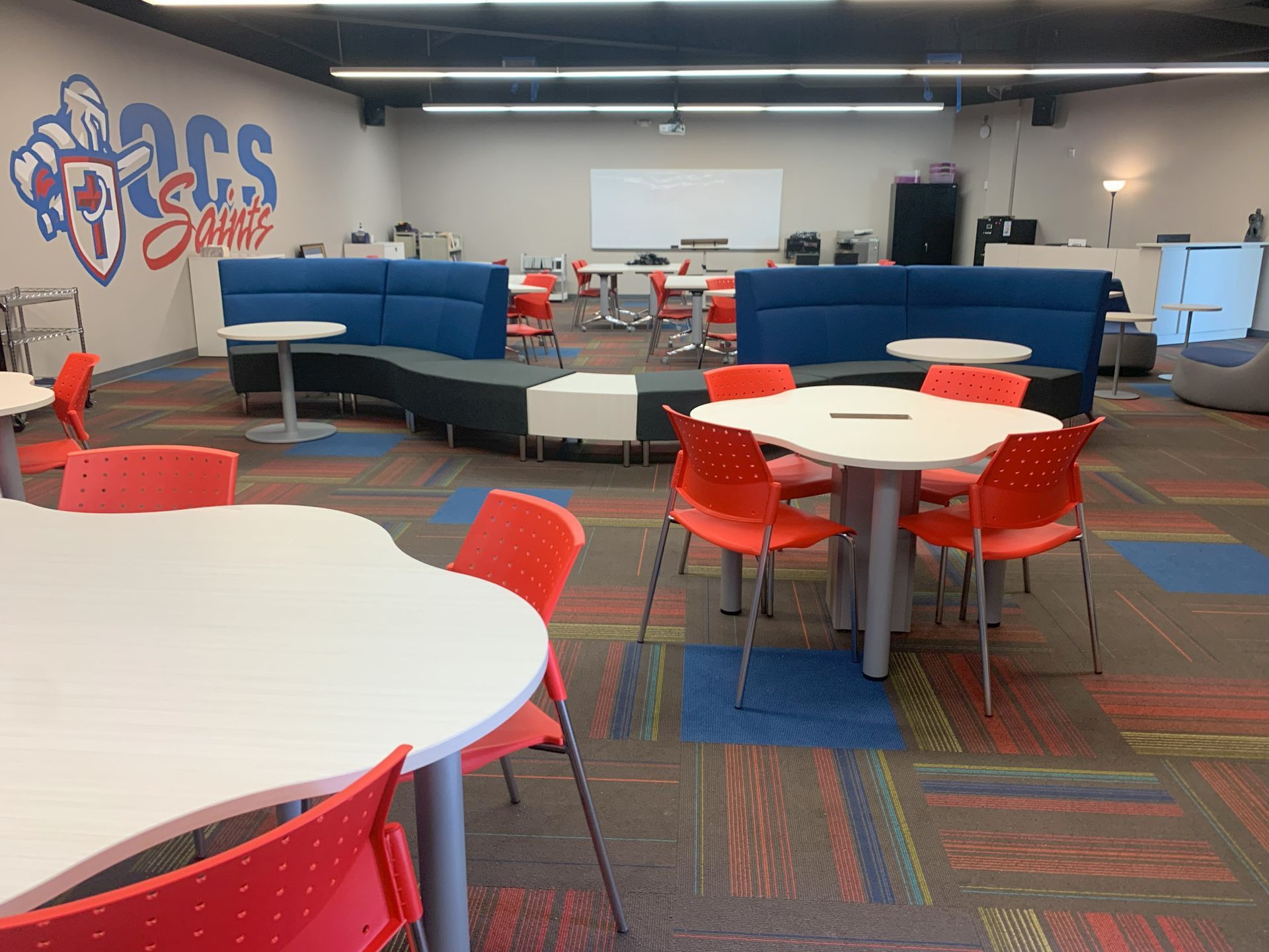 Elementary school media room with white tables and red chairs and a blue and gray sofa.