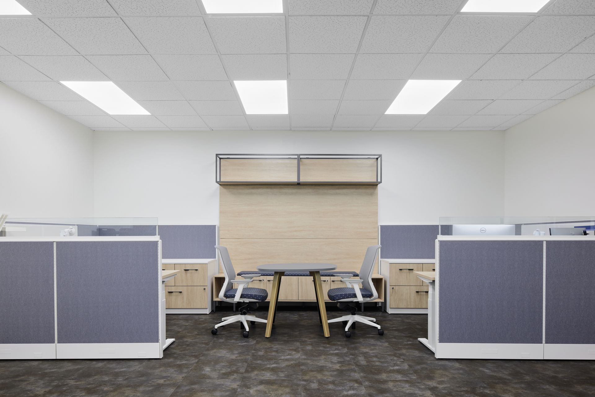 Office space with cubicles, two  chairs at a table, and light-colored overhead storage unit.