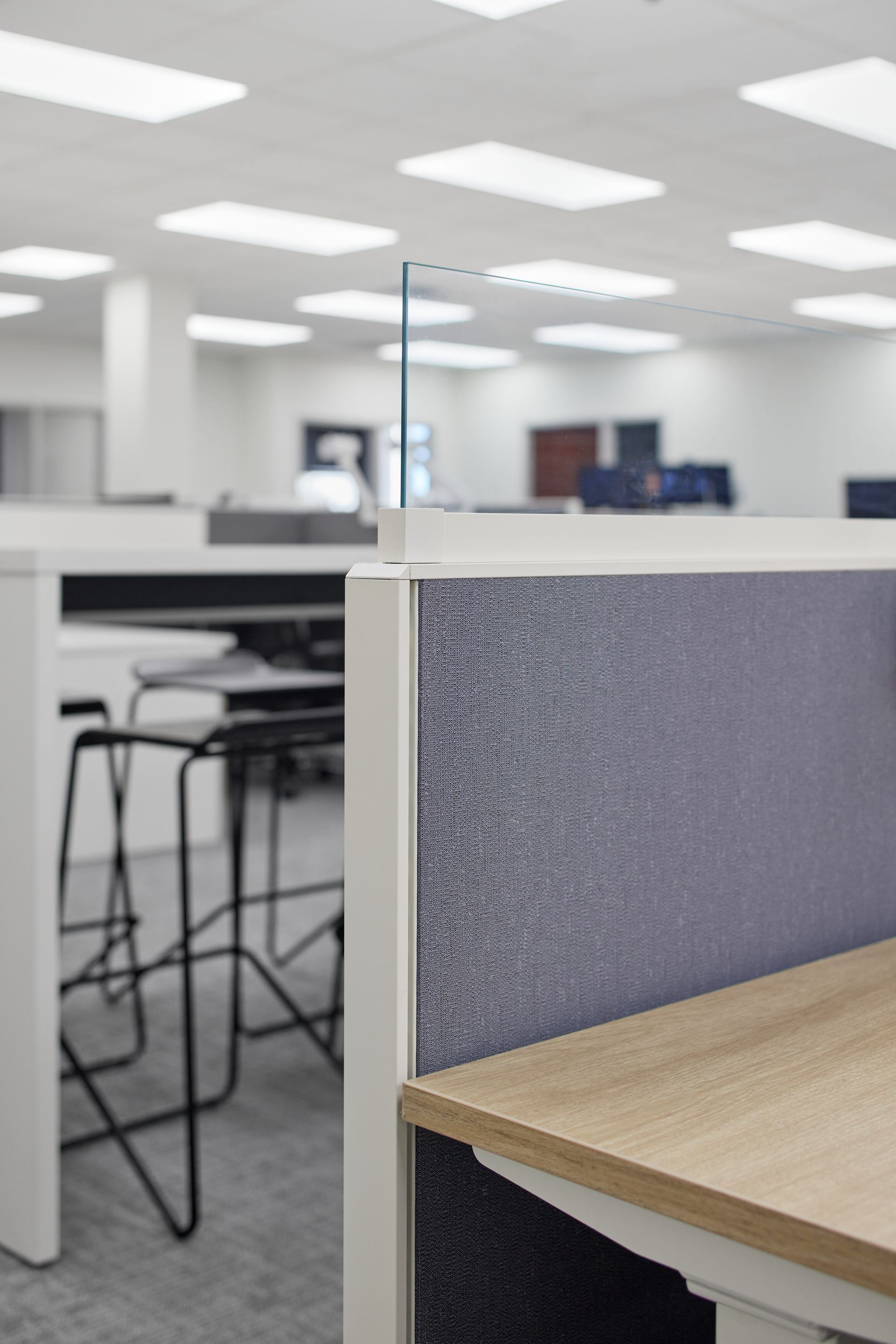 Detail view of a white framed cubicle panel with gray fabric and table with stools in background. 