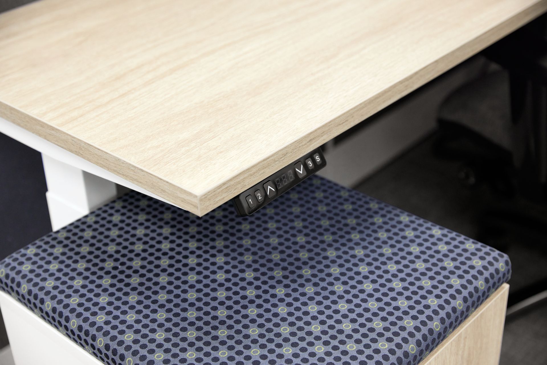 Height adjustable control panel for a light woodgrain desk above a mobile pedestal with navy circle patterned cushion.