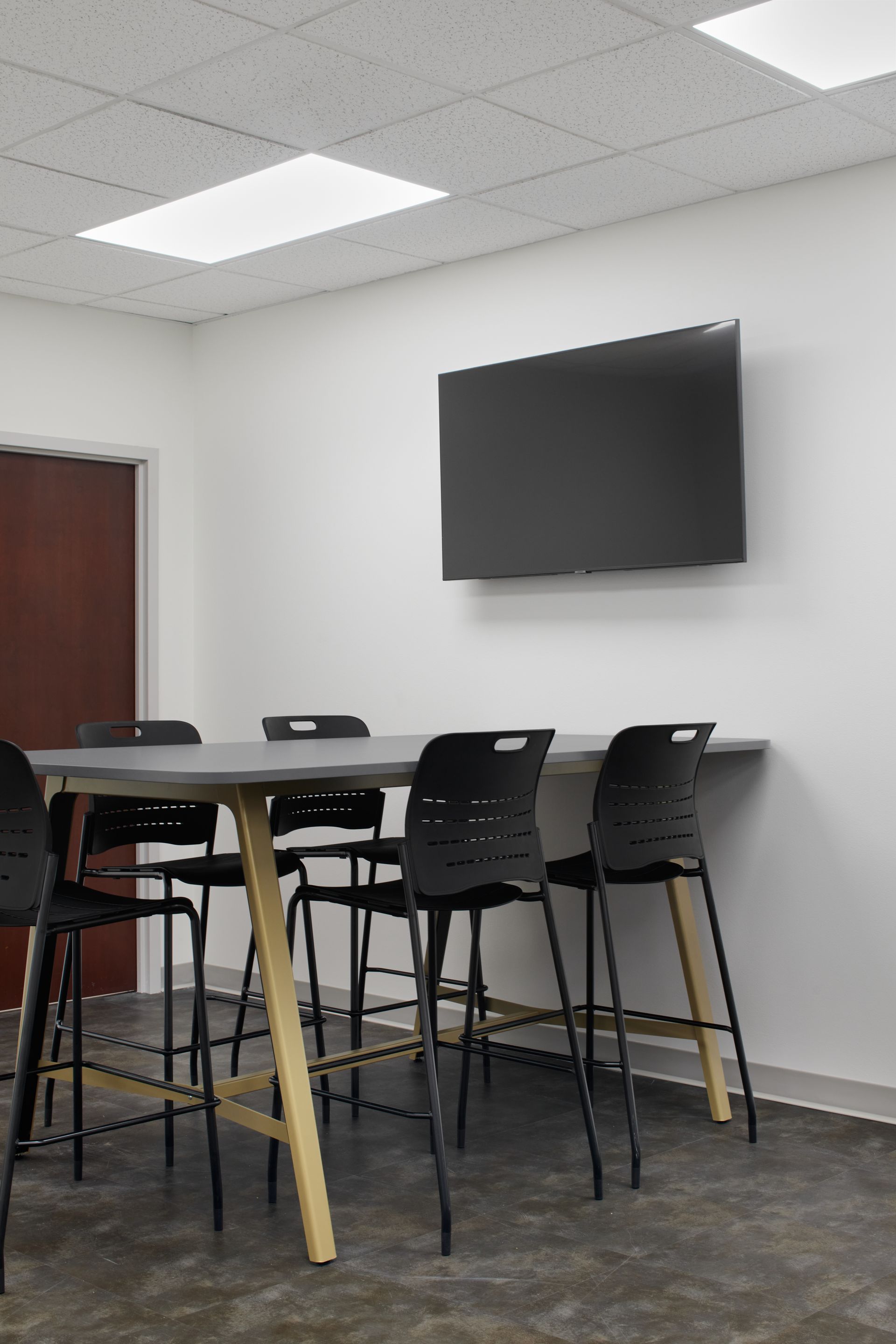 Gray meeting table with gold legs and five black stools sit against a light gray wall with TV above. 