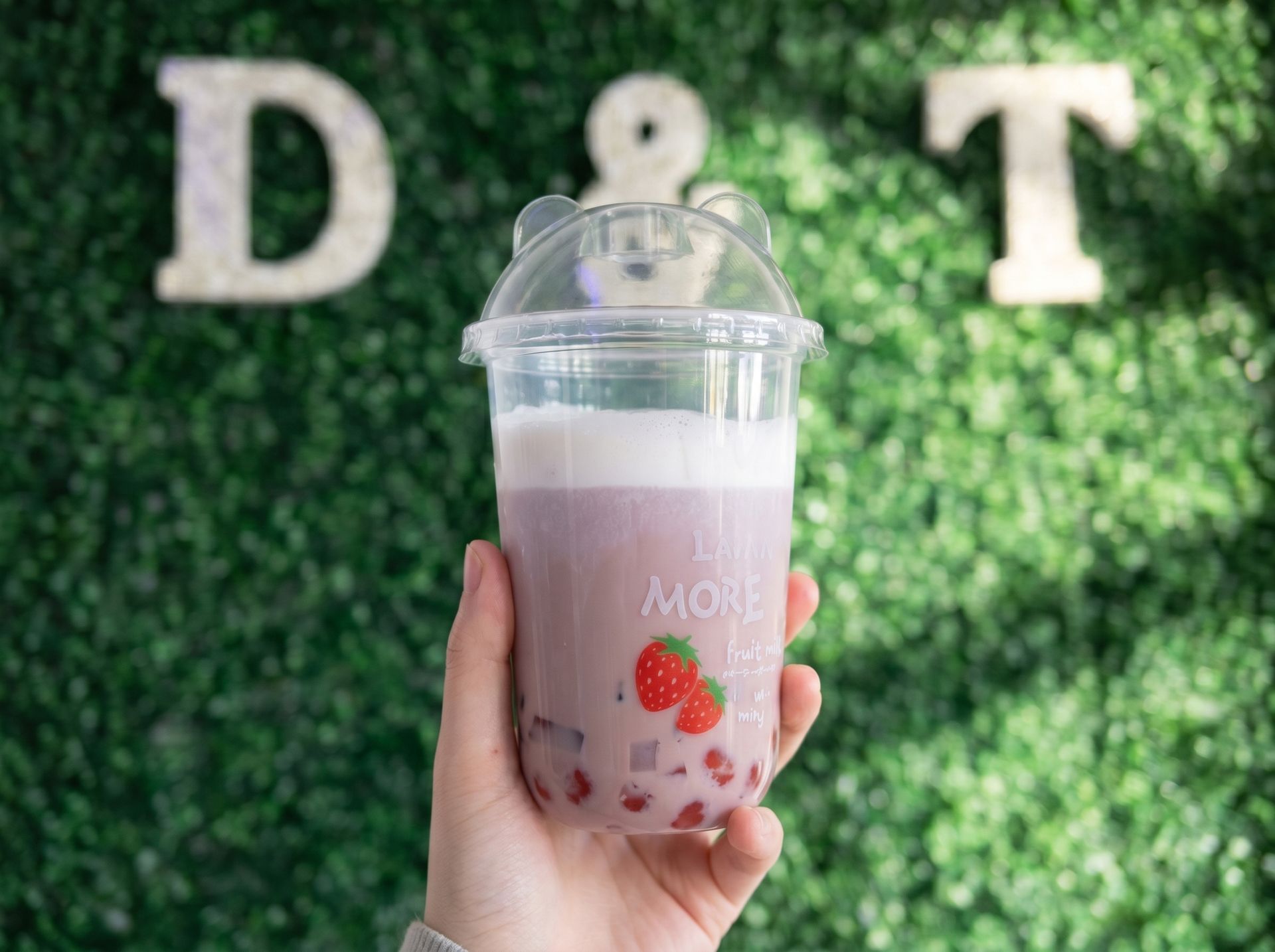 Hand holding a pink strawberry drink in a clear cup with lid, in front of a leafy wall with letters D and T.