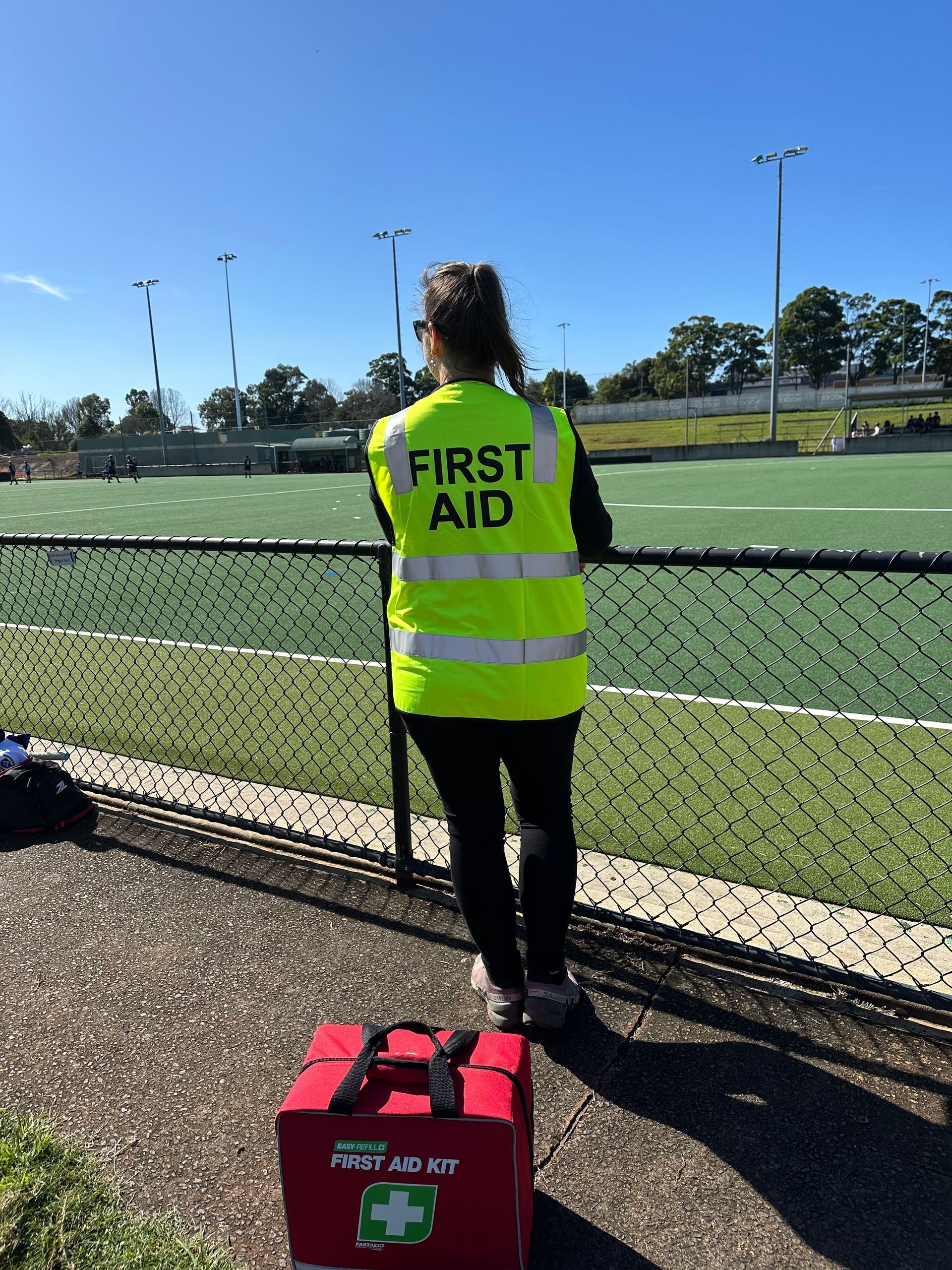 First Aid Worker in Yellow Vest Stands by a Chain-link Fence Beside a Sports Field — Revive Training in Alstonville, NSW