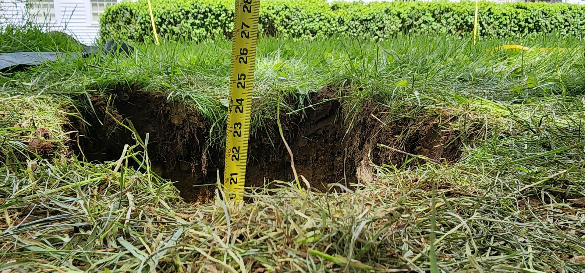 A freshly dug hole in grassy ground with a yellow measuring tape standing upright in the center.