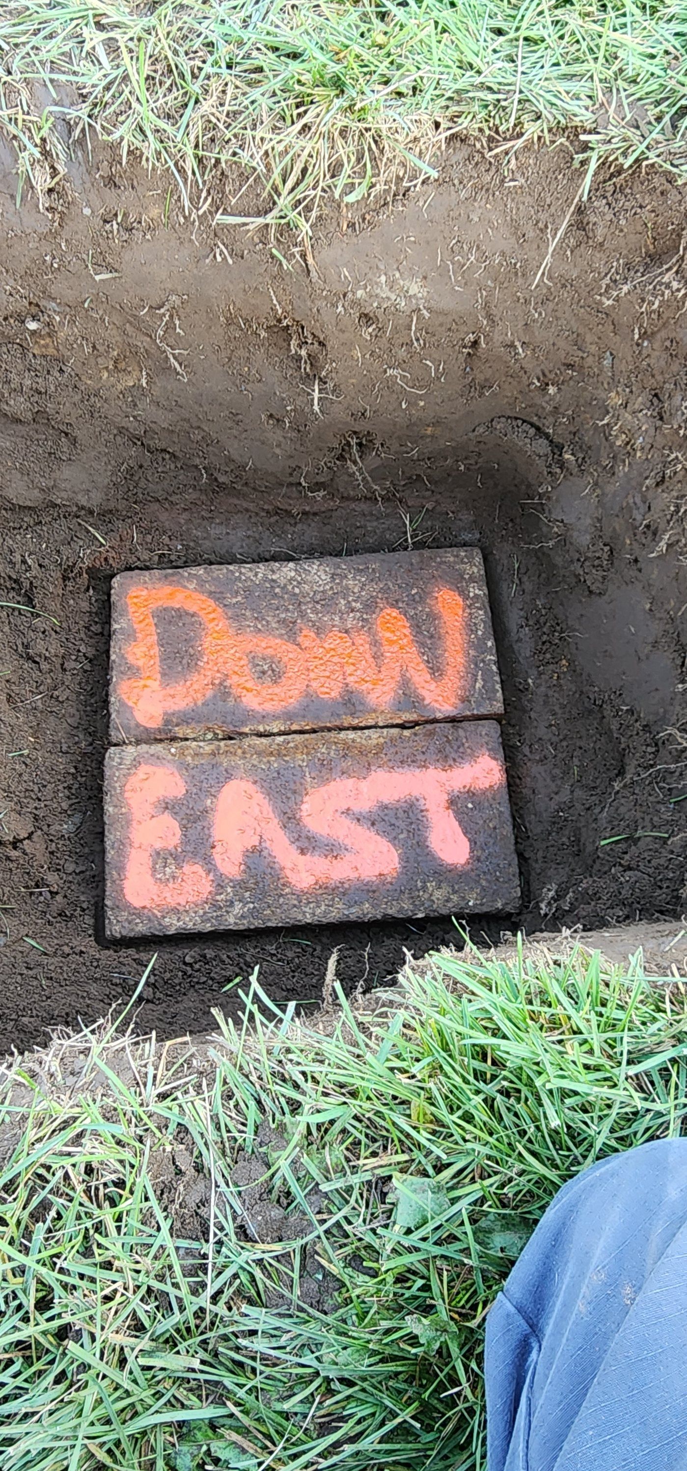 Orange “DOWN EAST” sign on dark soil beside grass outdoors