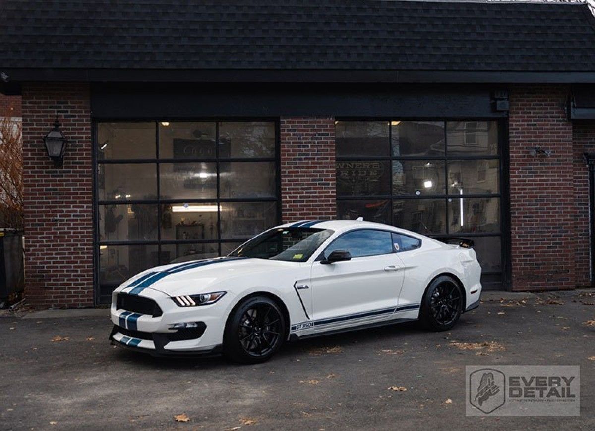 White Mustang with blue racing stripes parked in front of a brick garage