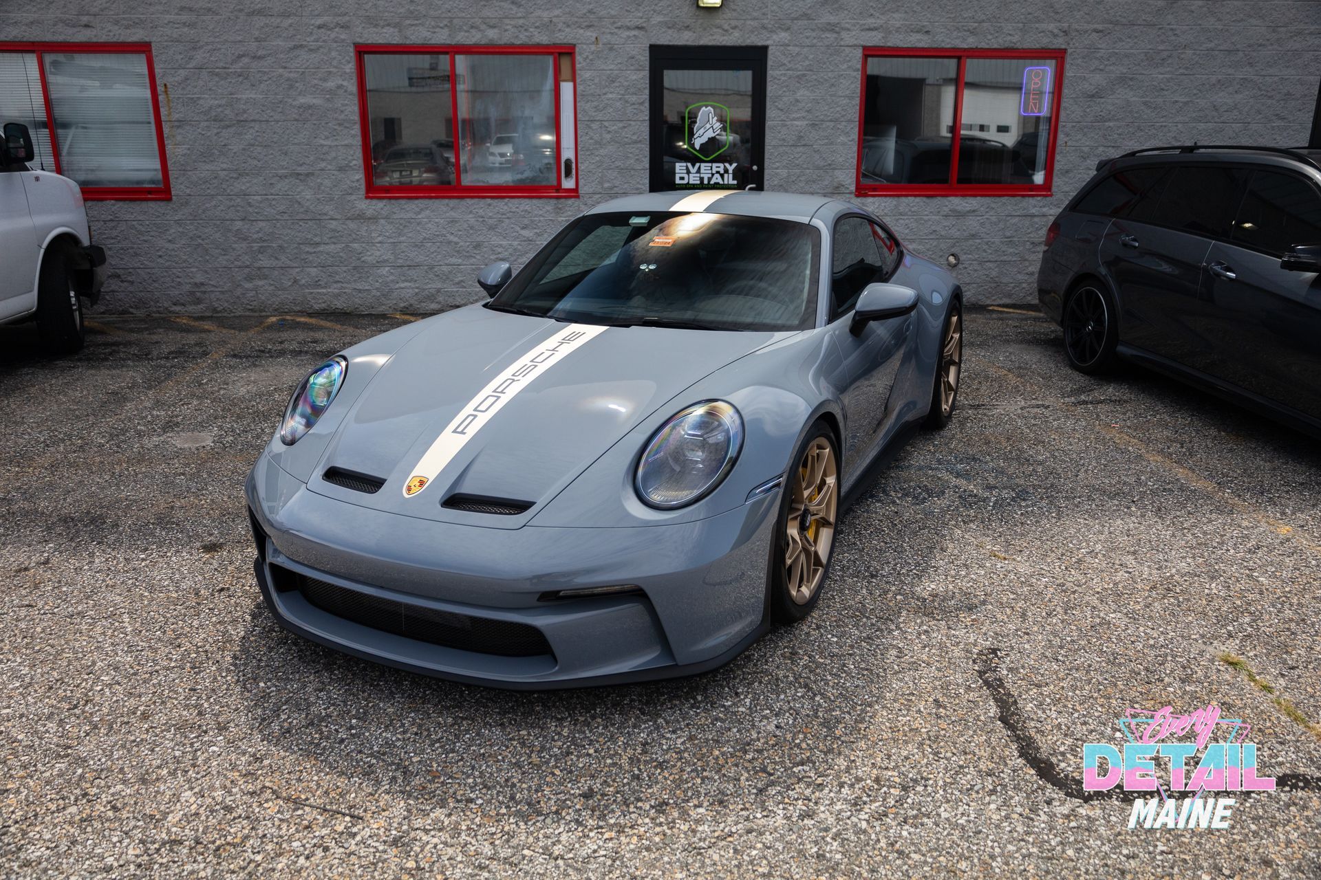 Gray Porsche 911 with white racing stripes parked outside a building with red-framed windows.