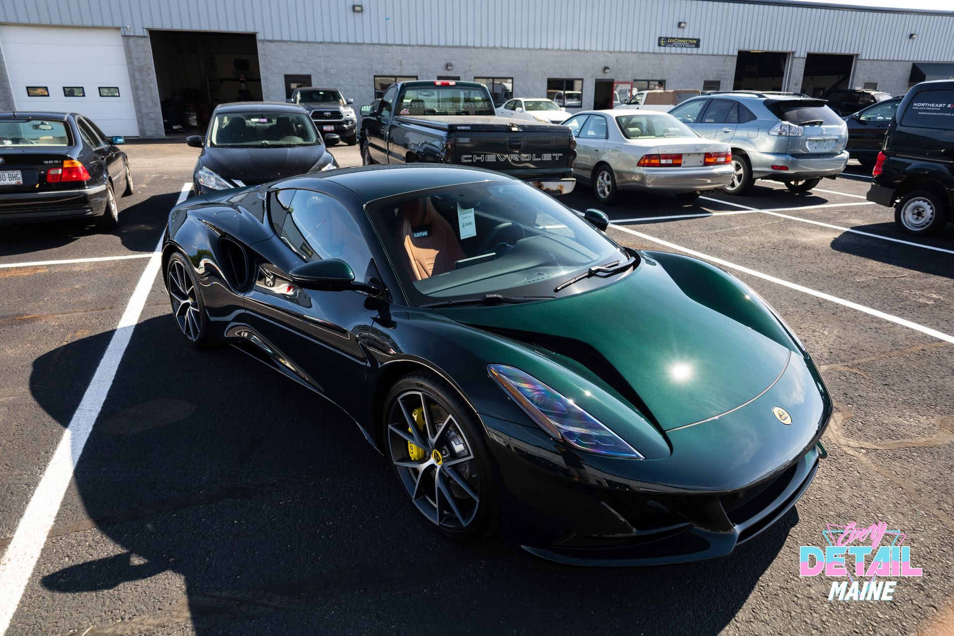 Dark green sports car parked in a lot with other cars and a warehouse building in the background.