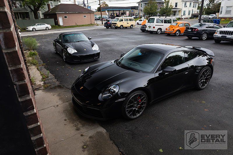 Two black Porsche sports cars parked on asphalt. Other cars and buildings in the background.