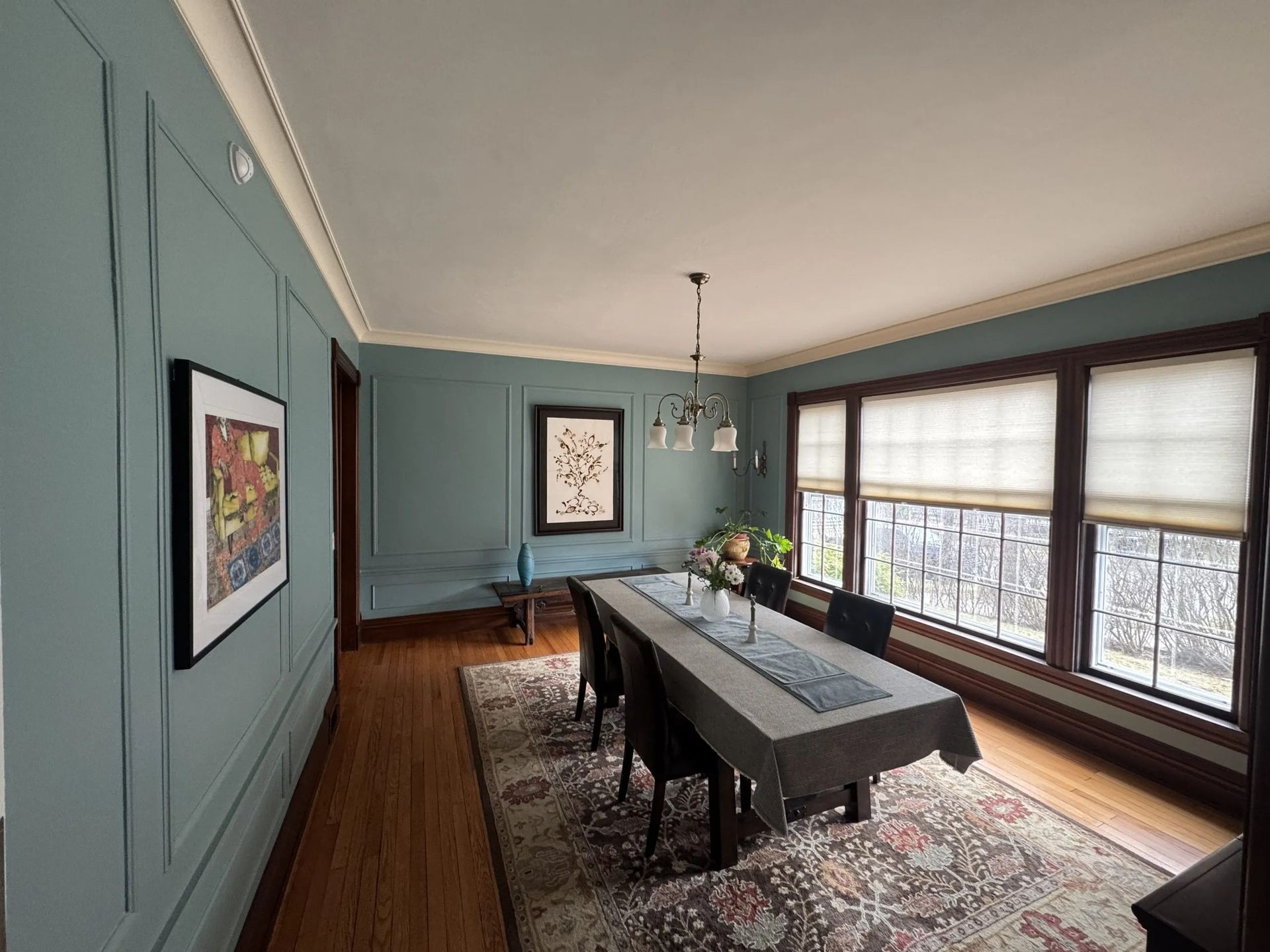 Dining room with blue walls, long table, and large windows with blinds overlooking a bright exterior.