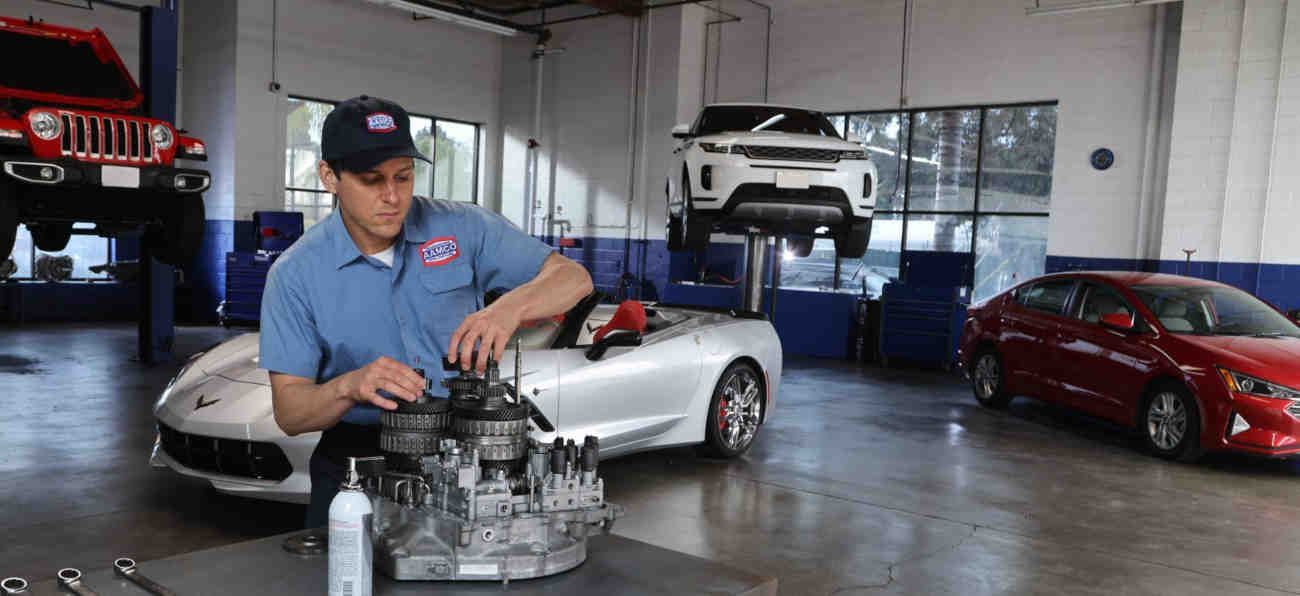 Mechanic in blue shirt working on a transmission in a garage with silver and red sports cars