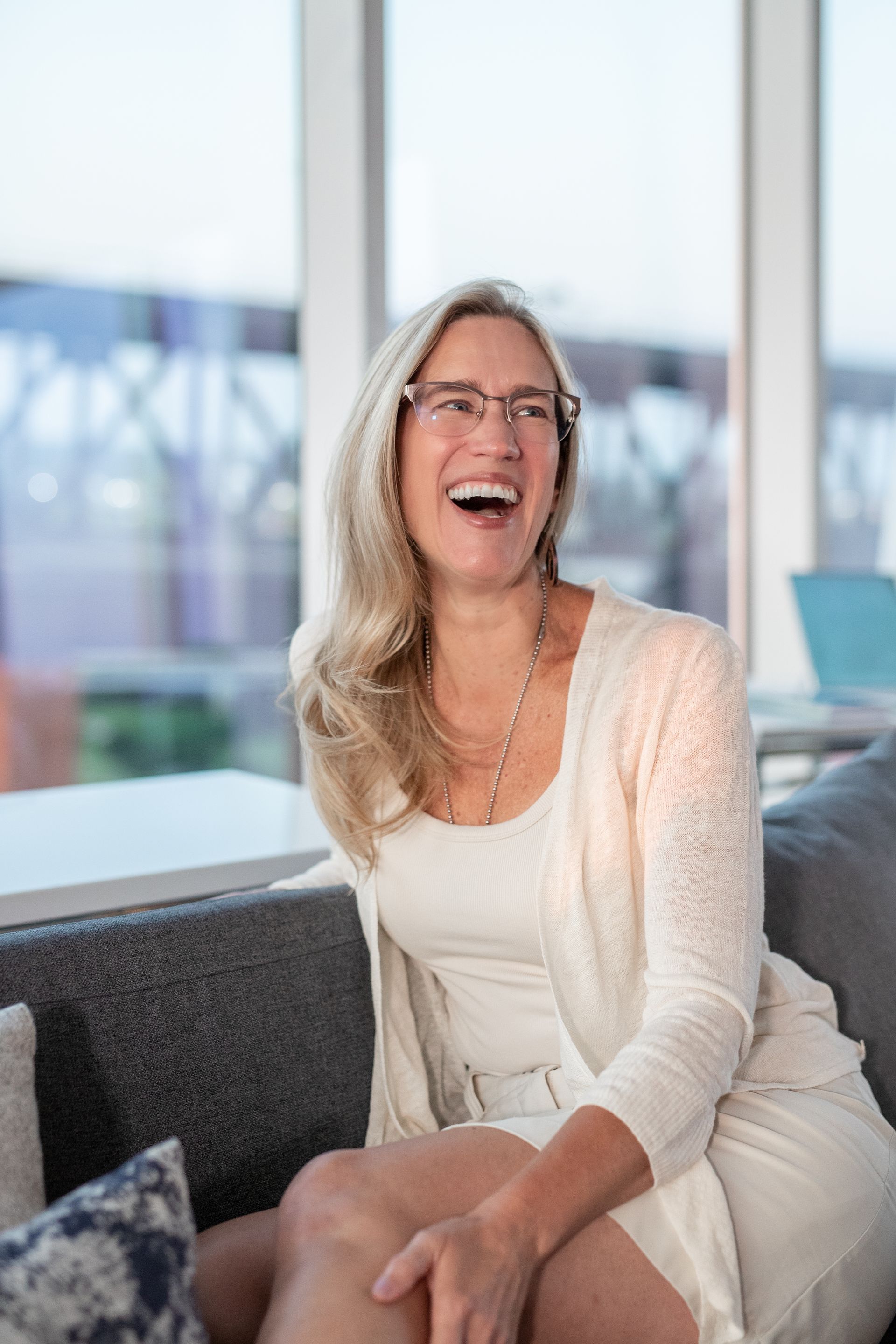 Woman in glasses laughing, seated on a gray couch in a bright room with a city view.