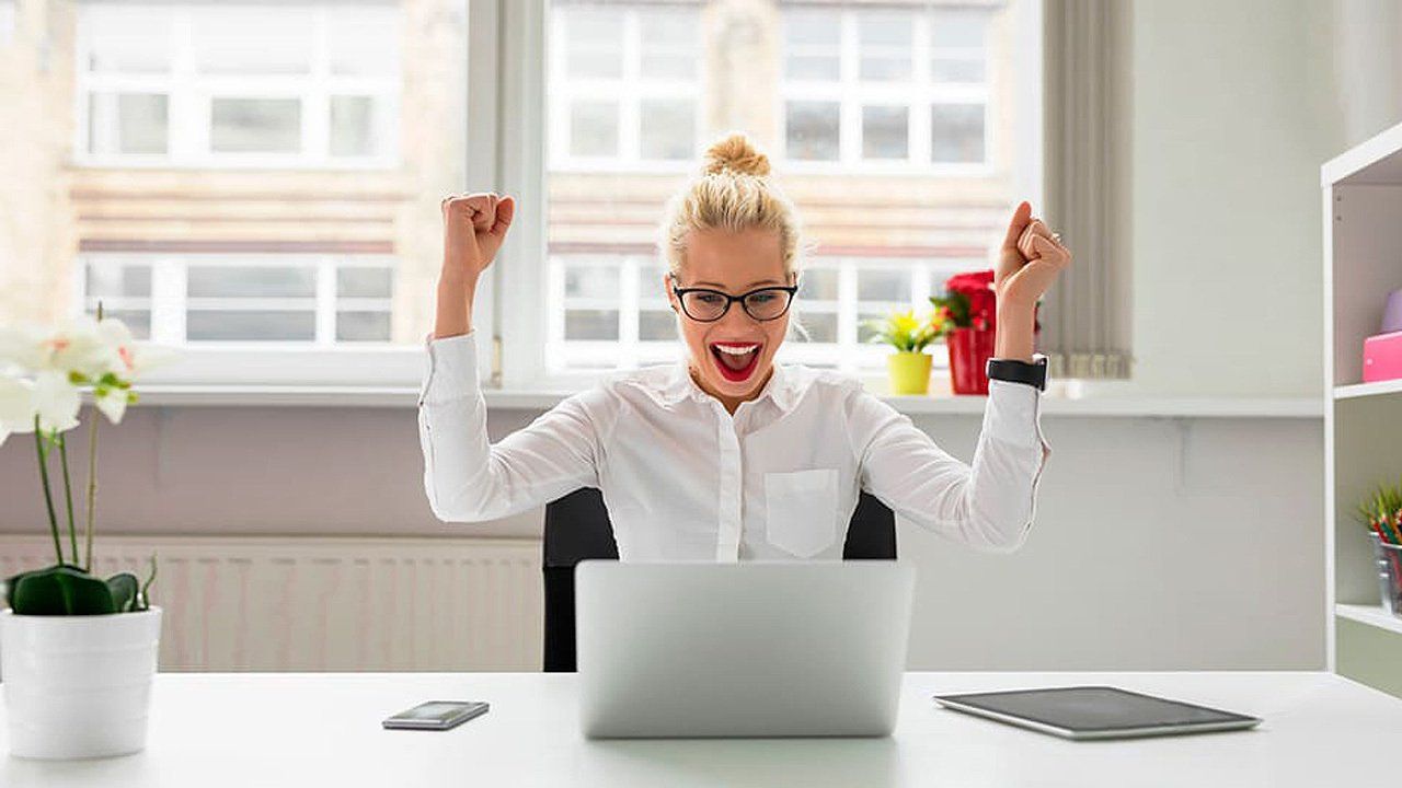 A woman is sitting at a desk in front of a laptop computer with her arms in the air.