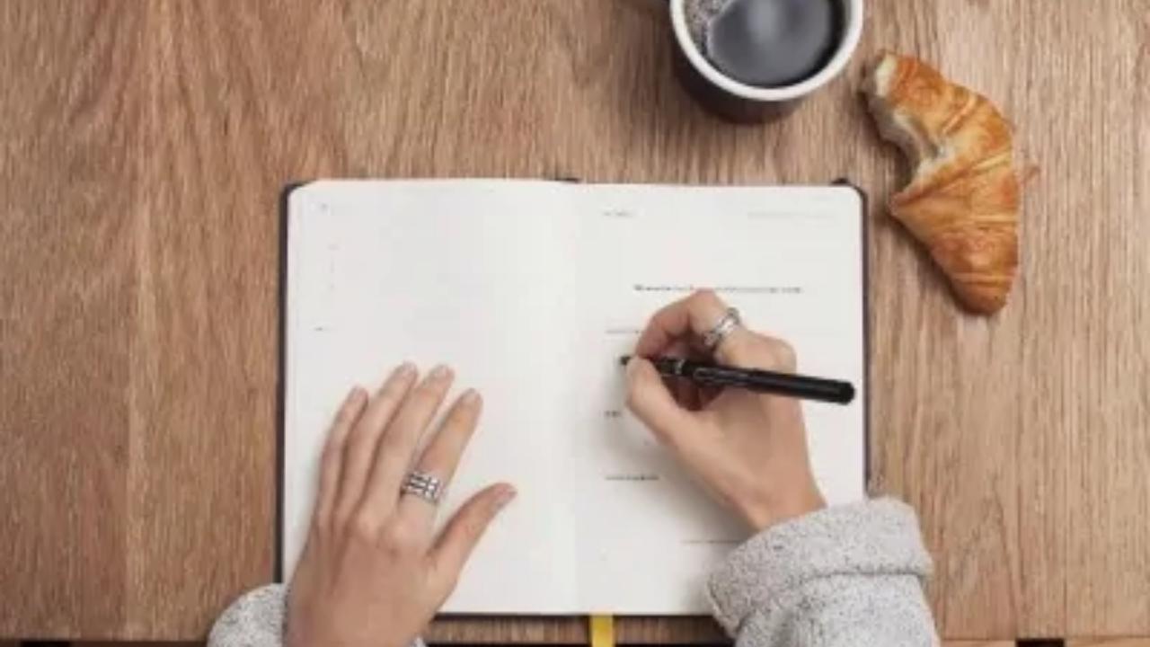 A person is writing in a notebook next to a cup of coffee and a croissant.