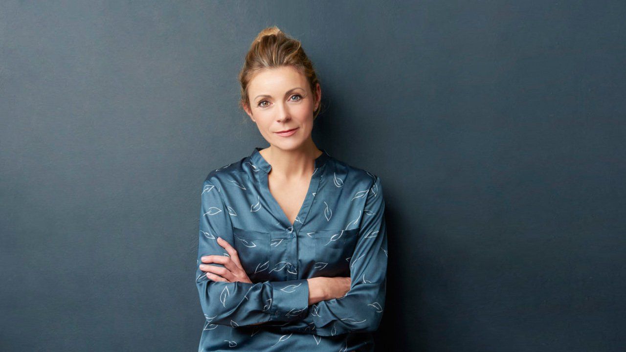 A woman in a blue shirt is standing with her arms crossed in front of a blue wall.