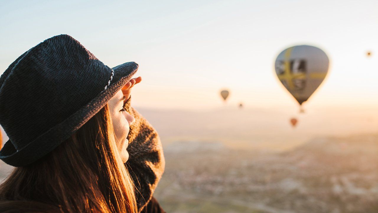 A woman in a hat is looking at hot air balloons in the sky.
