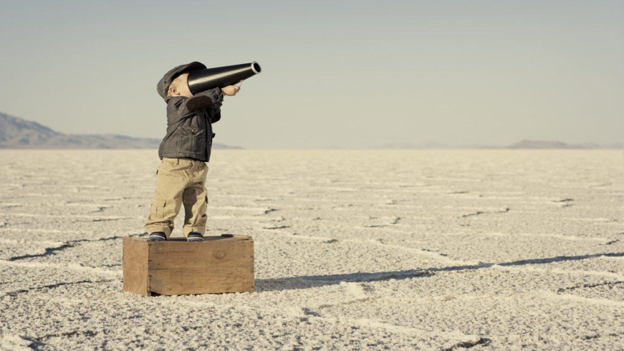 A young boy is standing on a box in the middle of a desert looking through binoculars.