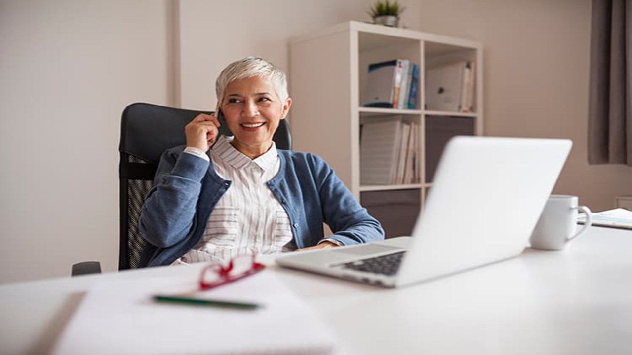 An older woman is sitting at a desk with a laptop and talking on a cell phone.