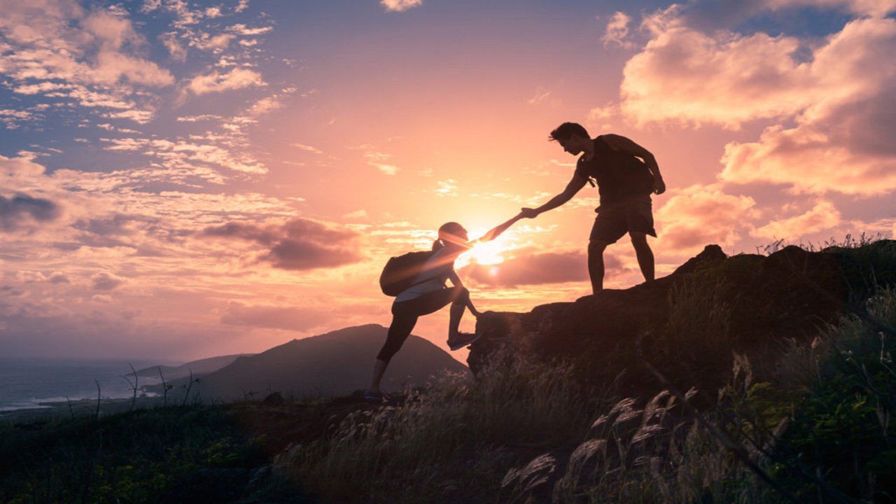 A man is helping another man climb a mountain at sunset.