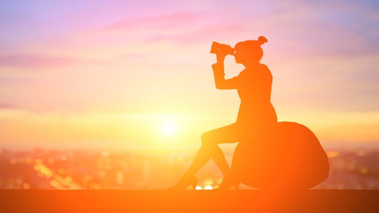 A woman is sitting on a suitcase looking through binoculars at sunset.