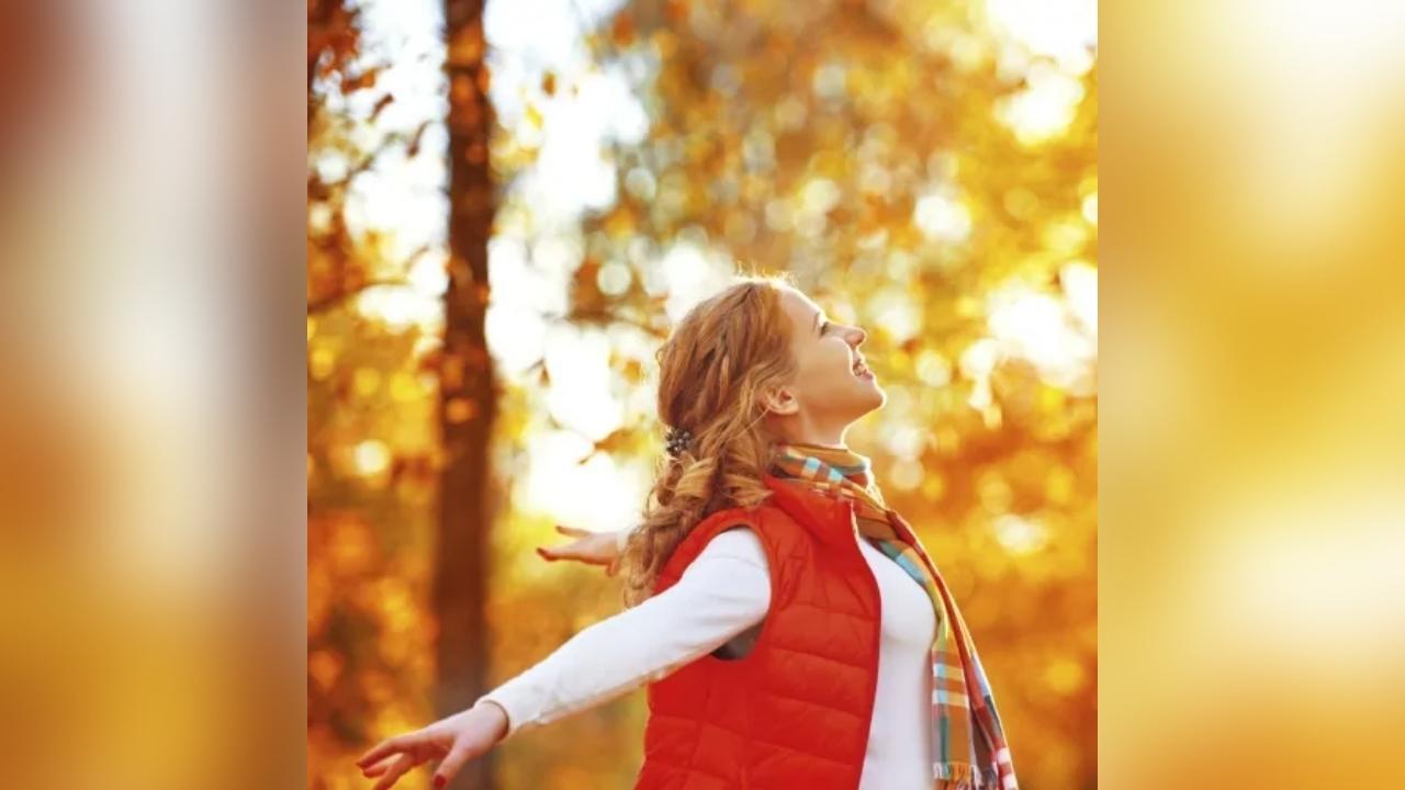 A woman in a red vest is standing in the woods with her arms outstretched.