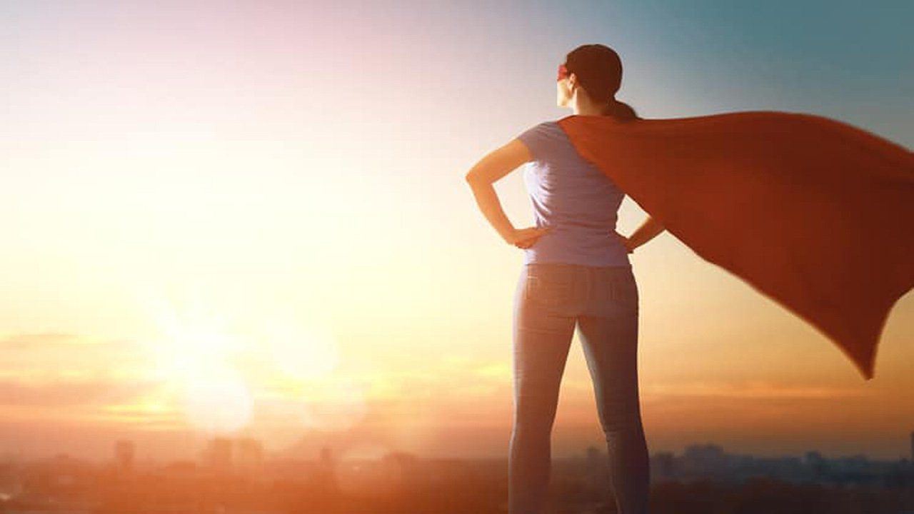 A woman in a superhero cape is standing in front of a city at sunset.
