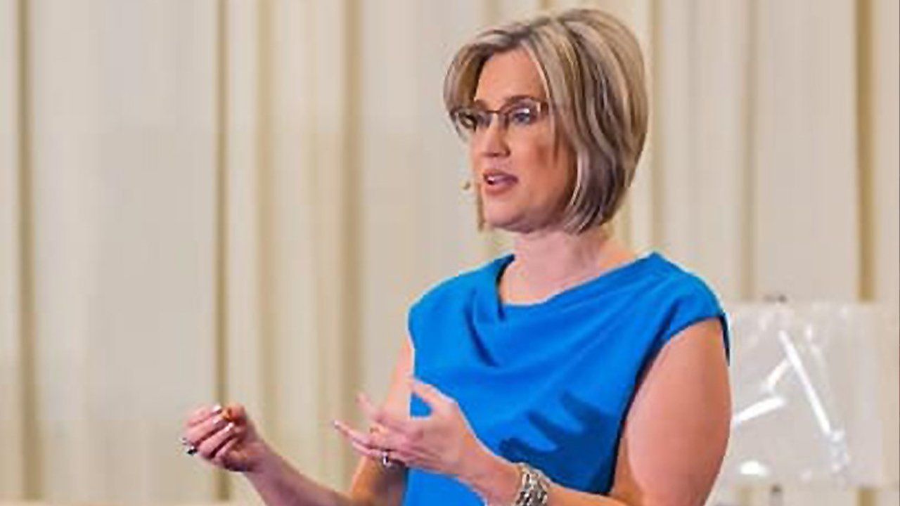 A woman in a blue dress and glasses is giving a speech.
