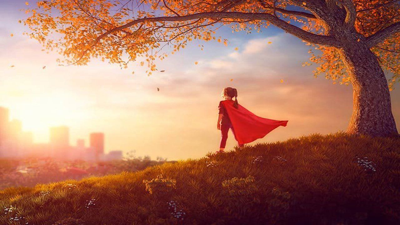 A little girl in a red cape is standing on a hill next to a tree.