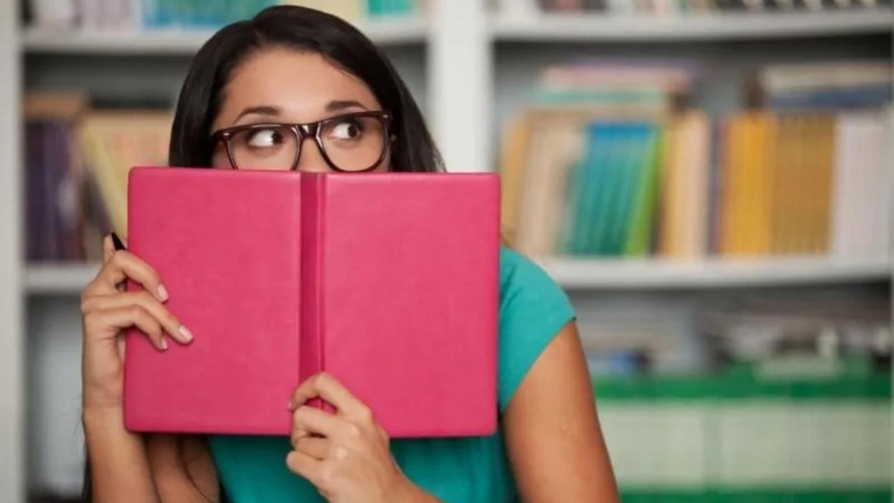 A woman is covering her face with a book in a library.