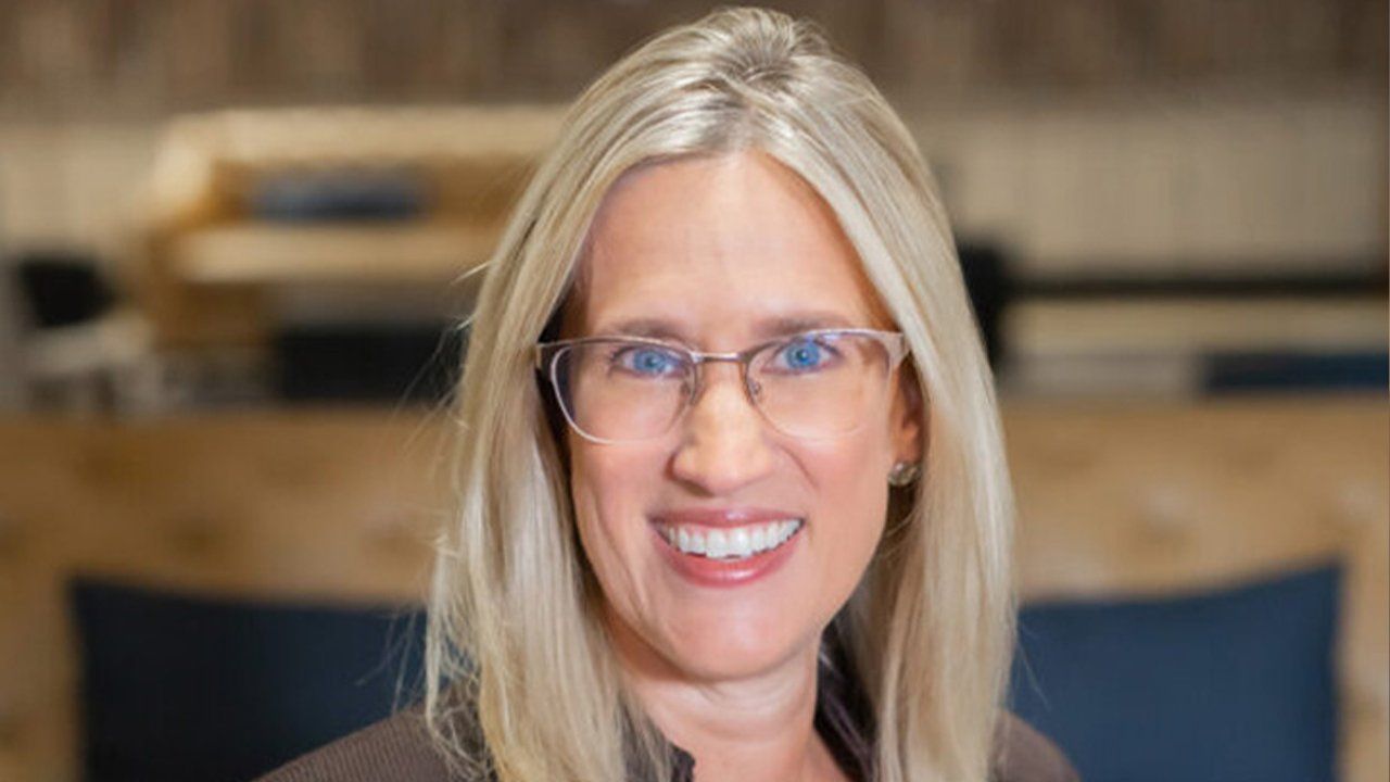 A woman wearing glasses and a brown shirt is smiling for the camera.