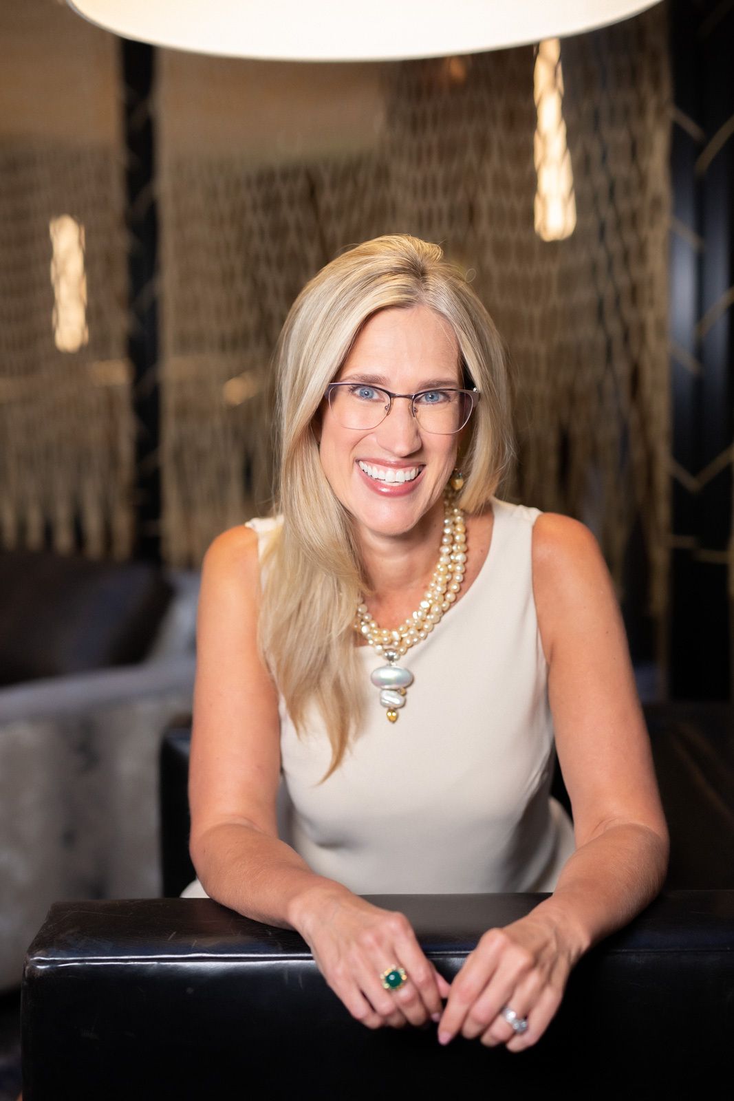 A woman wearing glasses and a necklace is sitting on a couch and smiling.
