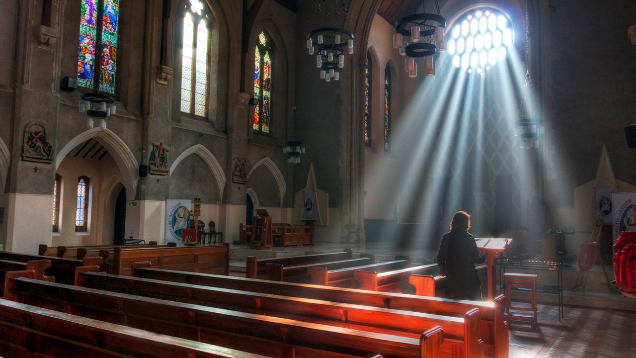 A person is praying in a church with the sun shining through the windows.