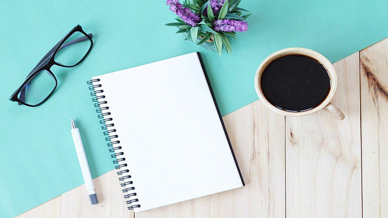 A notebook , pen , glasses , and a cup of coffee on a wooden table.
