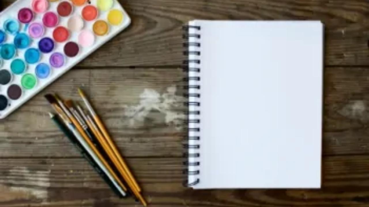 An empty notebook is sitting on a wooden table next to a palette of watercolors and brushes.