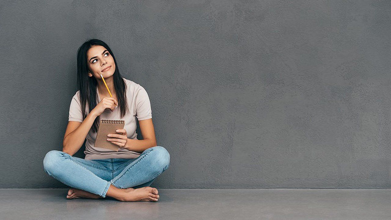 A woman is sitting on the floor with her legs crossed holding a notebook and a pencil.