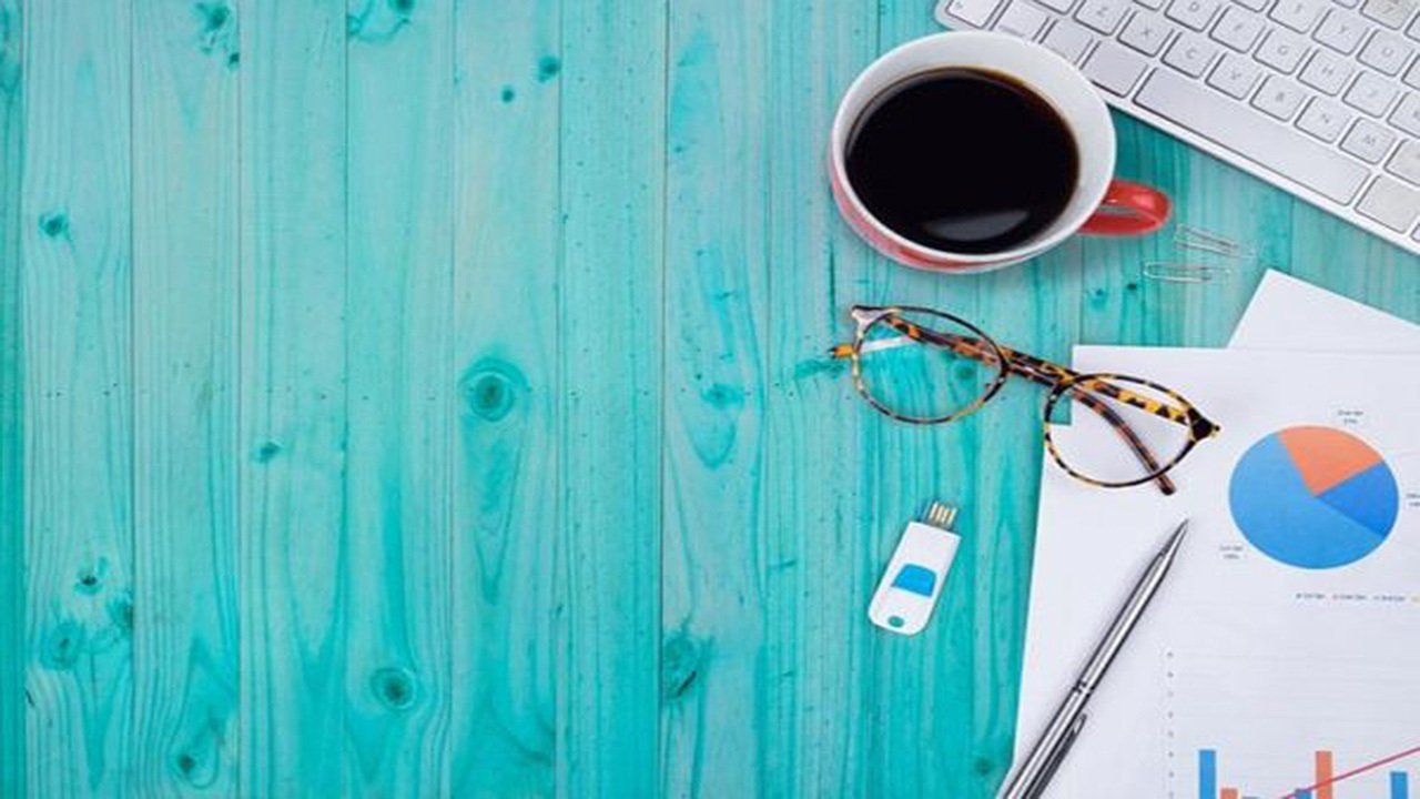 A wooden desk with a cup of coffee , glasses , a pen , a keyboard , and a usb drive.