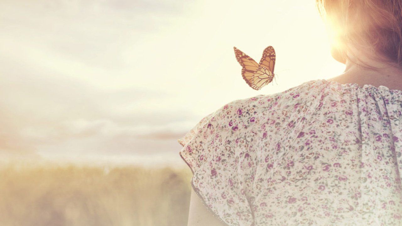 A woman is standing in a field with a butterfly on her shoulder.