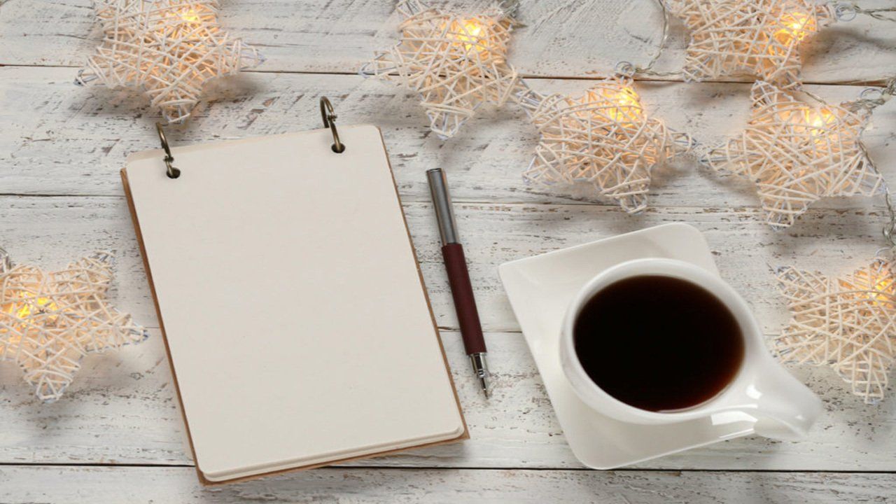 A cup of coffee , a notebook , a pen and christmas lights on a wooden table.