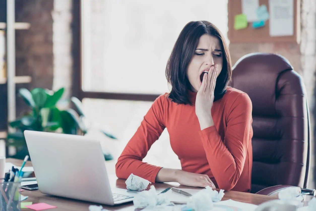 A woman is yawning while sitting at a desk with a laptop.