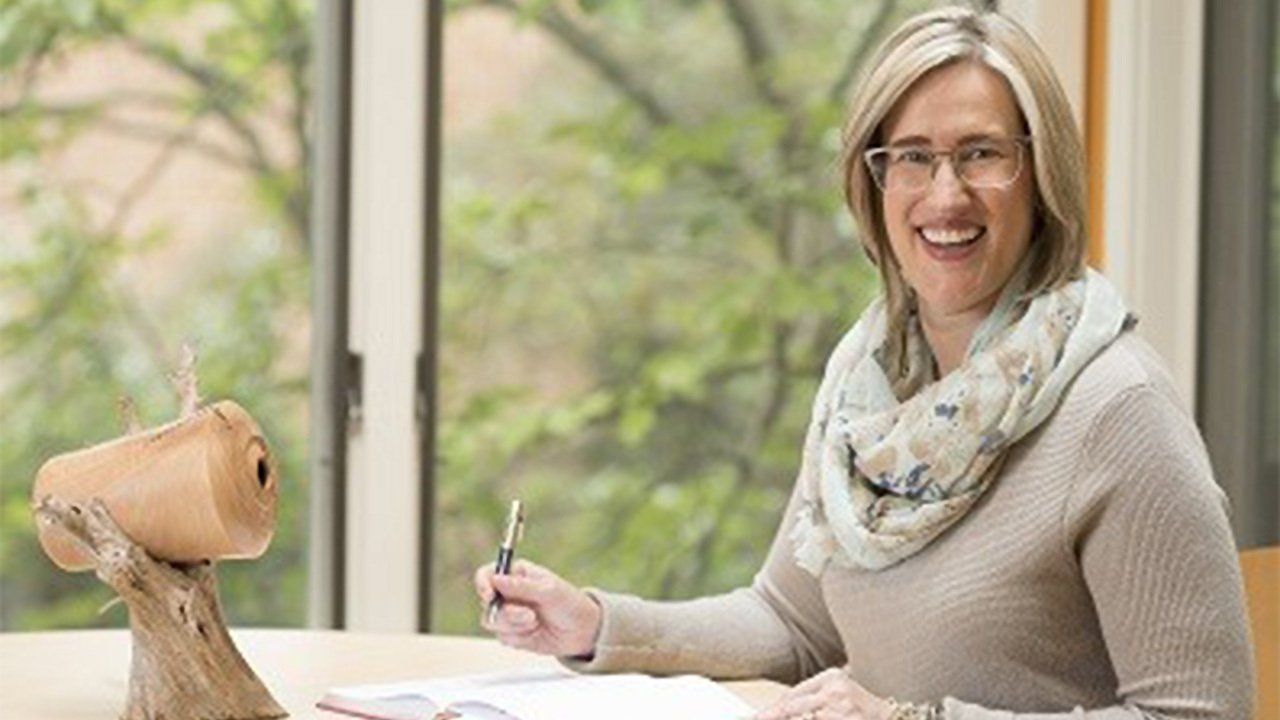 A woman is sitting at a desk in front of a window holding a pen.