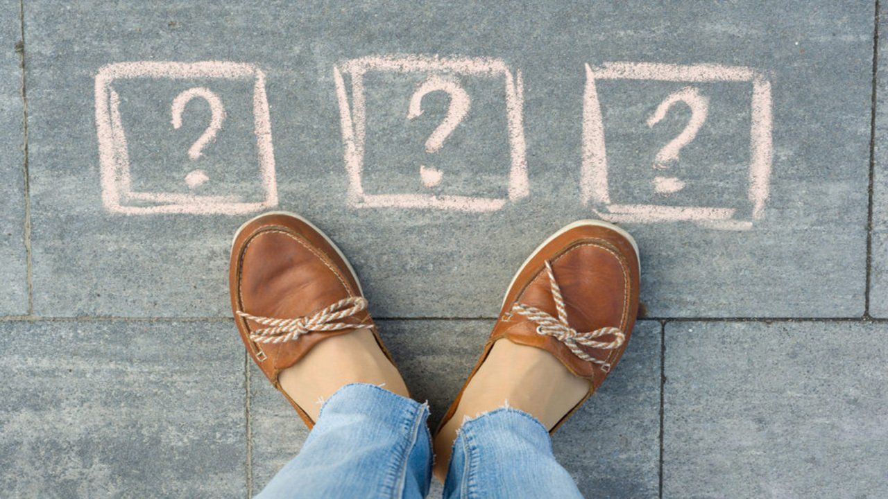 A person is standing in front of a chalkboard with three question marks drawn on it.
