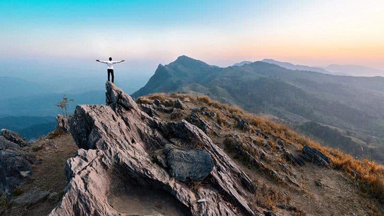 A man is standing on top of a rocky mountain with his arms outstretched.