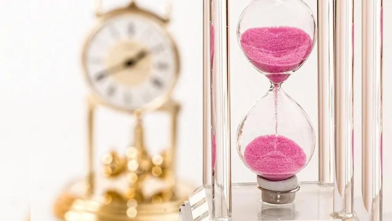 A close up of a hourglass with pink sand and a clock in the background.