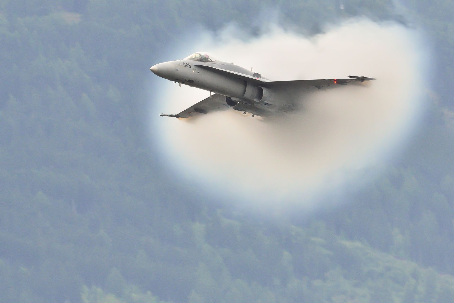 Fighter jet traveling fast, creating a vapor cone around it, set against a backdrop of green mountains and sky.
